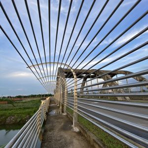 A modern pedestrian bridge featuring metallic railings creates a series of parallel lines that lead the viewer's eye towards a blue sky with wispy clouds. 