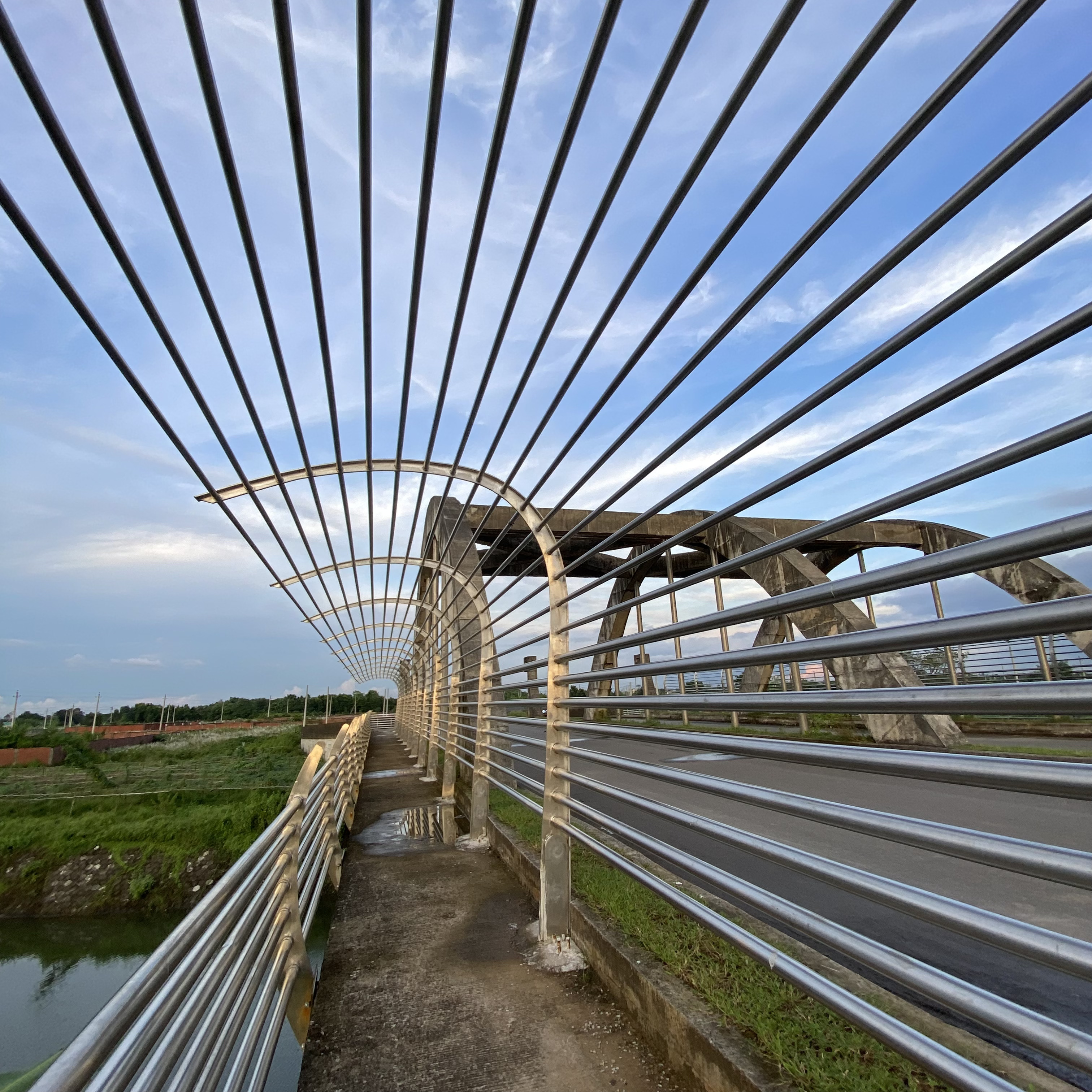 A modern pedestrian bridge featuring metallic railings creates a series of parallel lines that lead the viewer's eye towards a blue sky with wispy clouds.