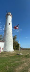 A lighthouse tower on a green lawn with a blue sky, next to a flag pole with an American flag. Sturgeon Point Lighthouse, Michigan.