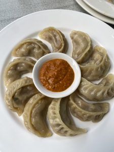 A plate featuring a circular arrangement of steamed dumplings, typically known as momos, surrounding a small white bowl filled with orange dipping sauce. 