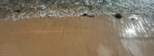 A low wave washing up on sand.  Seen from above, the water is at the top of the image, wet flat sand at the bottom.