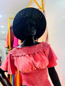 A black mannequin wears a pink off-the-shoulder top with a smocked bodice and a wide-brimmed black hat in a store display.