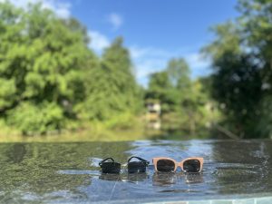 Two pairs of sunglasses, resting on the wet, black stone edge of an infinity pool, with a lush green forest and bright blue sky blurred in the background.