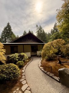 A curved, stone-lined path leads to a traditional Japanese tea house, set beneath a cloudy sky and the sun, in the Portland Japanese Garden of Oregon.