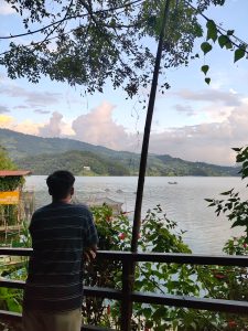 A person stands by a railing, overlooking a serene lake with hills, a drifting boat, red flowers, and a nearby lodge sign.