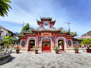 The pink Fujian Assembly Hall in Hoi An Ancient Town, Vietnam, with ornate green-tiled roofs, red Chinese characters above the entrance, and potted plants arranged in front under a bright blue sky.