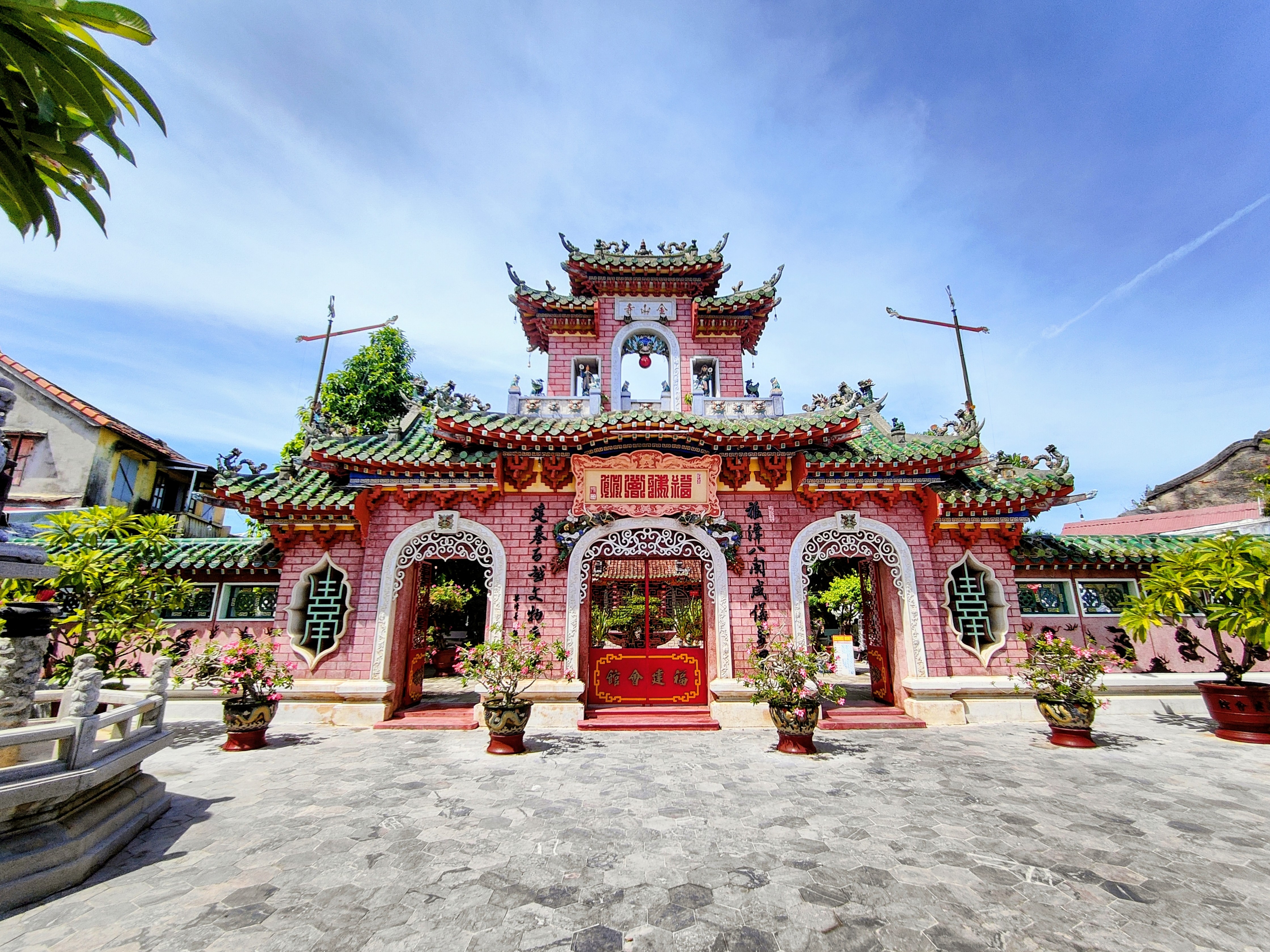 The pink Fujian Assembly Hall in Hoi An Ancient Town, Vietnam, with ornate green-tiled roofs, red Chinese characters above the entrance, and potted plants arranged in front under a bright blue sky.