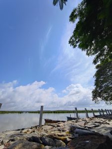 A riverside scene in Kundapura with small fishing boats tied near the shore, rocks in the foreground, and a bright blue sky with scattered clouds. Green trees hang from the top right corner.