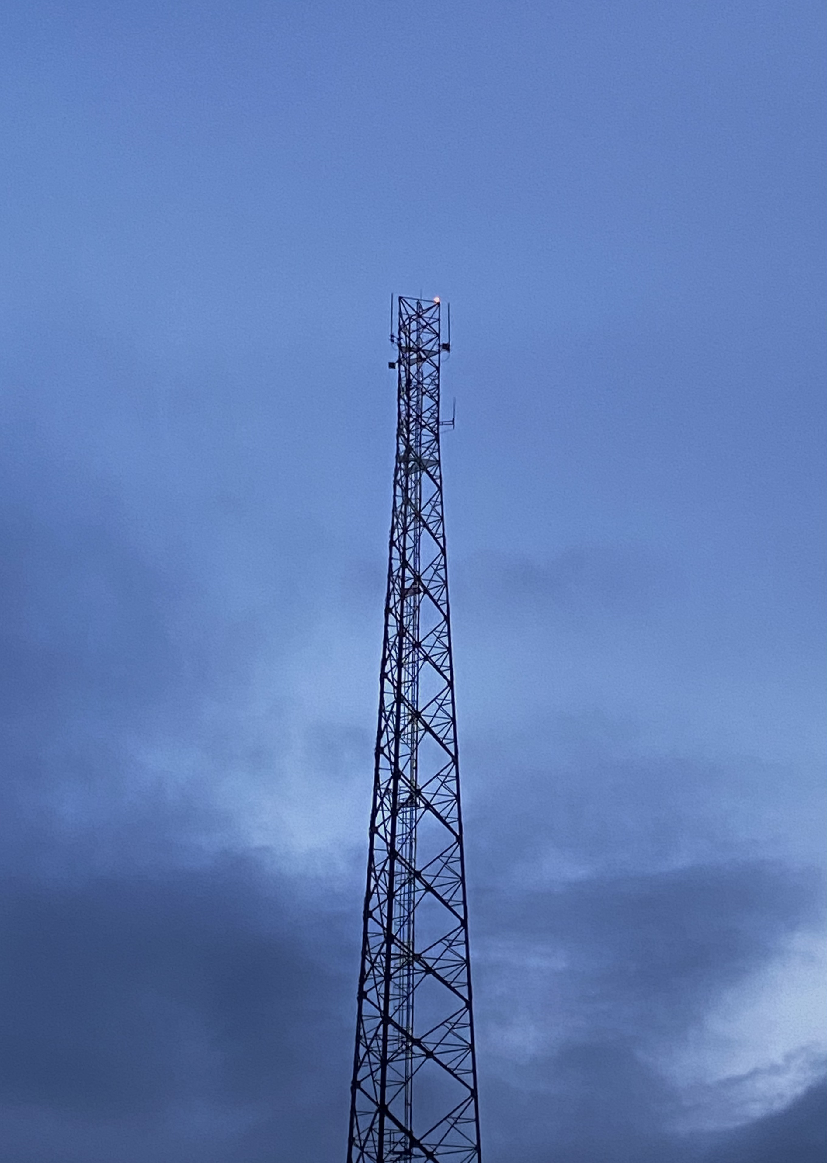 A tall, metal communication tower rises into a cloudy, twilight sky. The tower is constructed with a lattice design and has antennas mounted at the top and sides