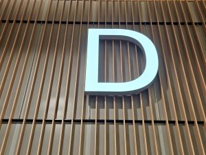 A large white letter &quot;D&quot; sign mounted on a wall with vertical wooden slats. Captured inside Portland International Airport, this photo shows the minimalist design used for gate or section identifiers in the terminal. 