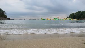 Cloudy beach with gentle waves, inflatable water attractions, and greenery along the shore.