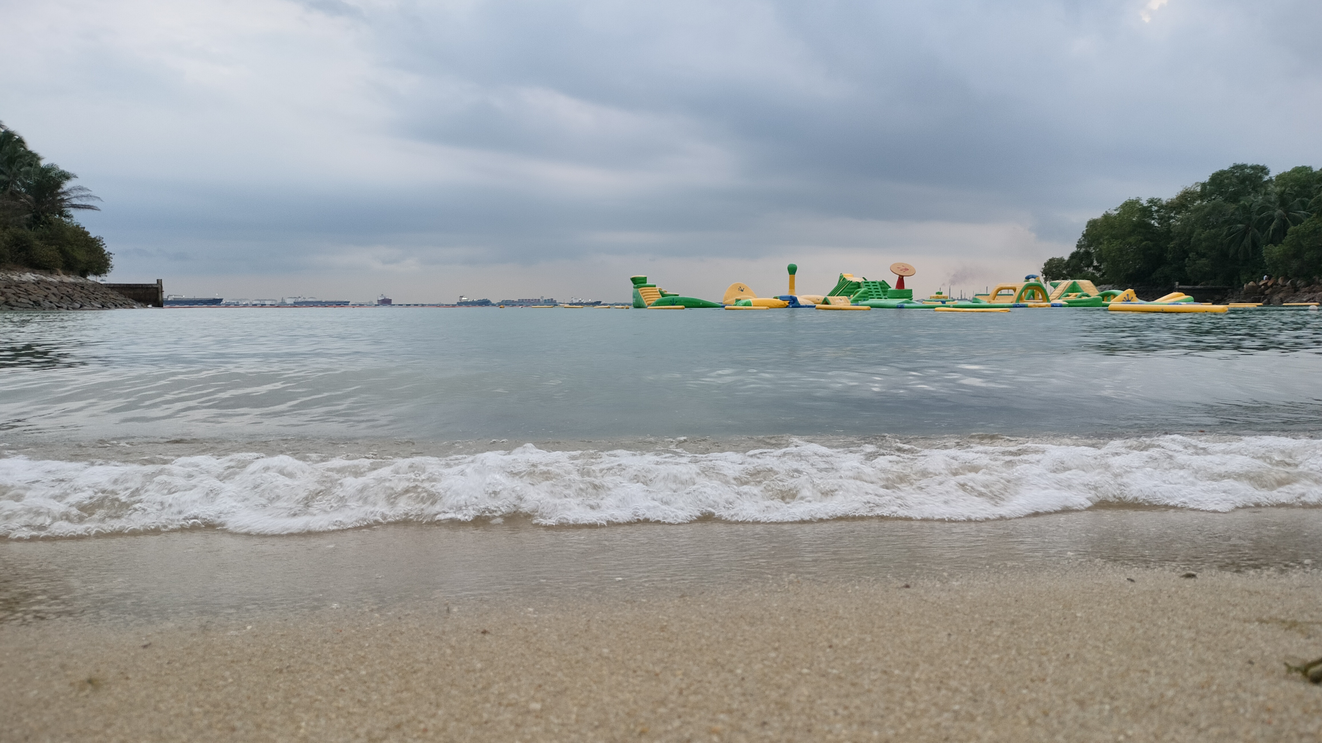 Cloudy beach with gentle waves, inflatable water attractions, and greenery along the shore.