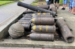 A display of antique, rusty artillery shells, including a large cannon and a round cannonball, sits on a concrete slab outdoors.