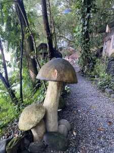 A large decorative mushroom sculpture stands beside a pebble pathway surrounded by lush greenery. 