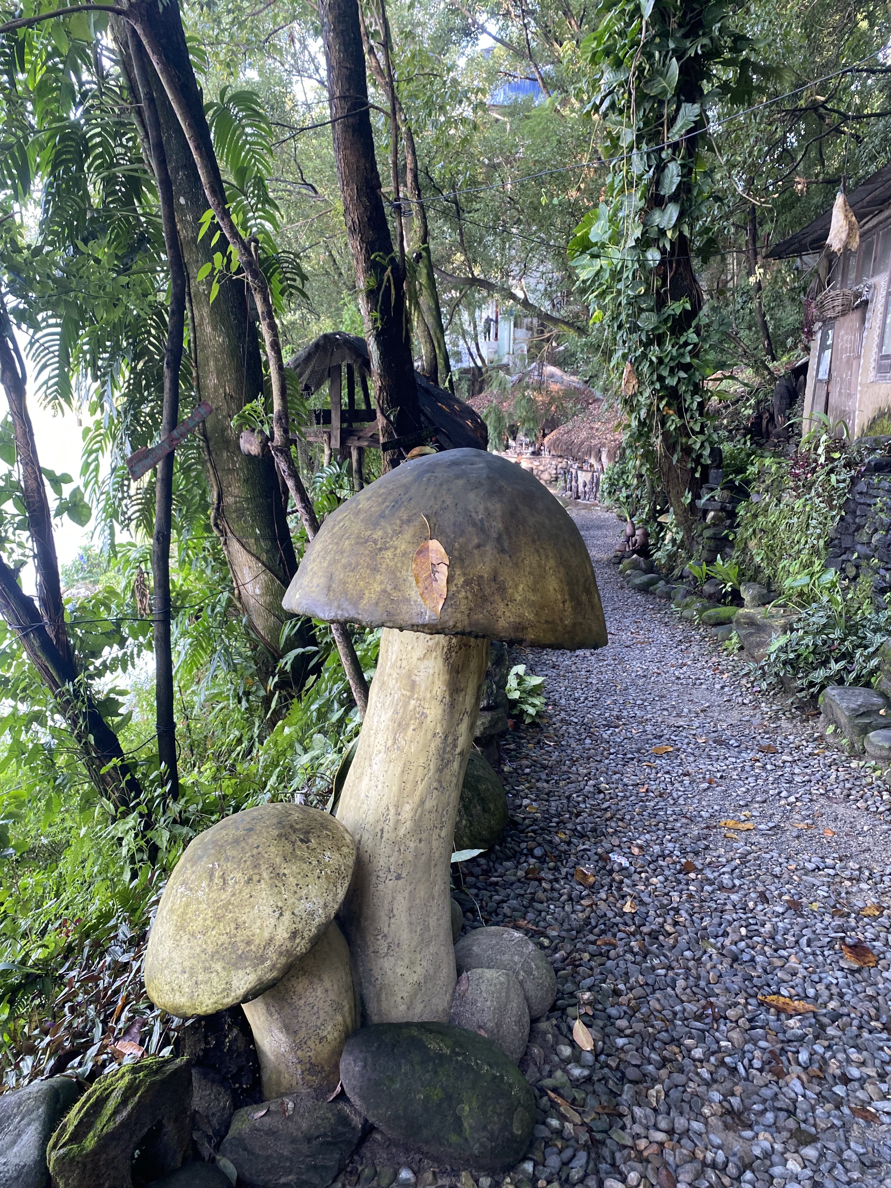 A large decorative mushroom sculpture stands beside a pebble pathway surrounded by lush greenery.