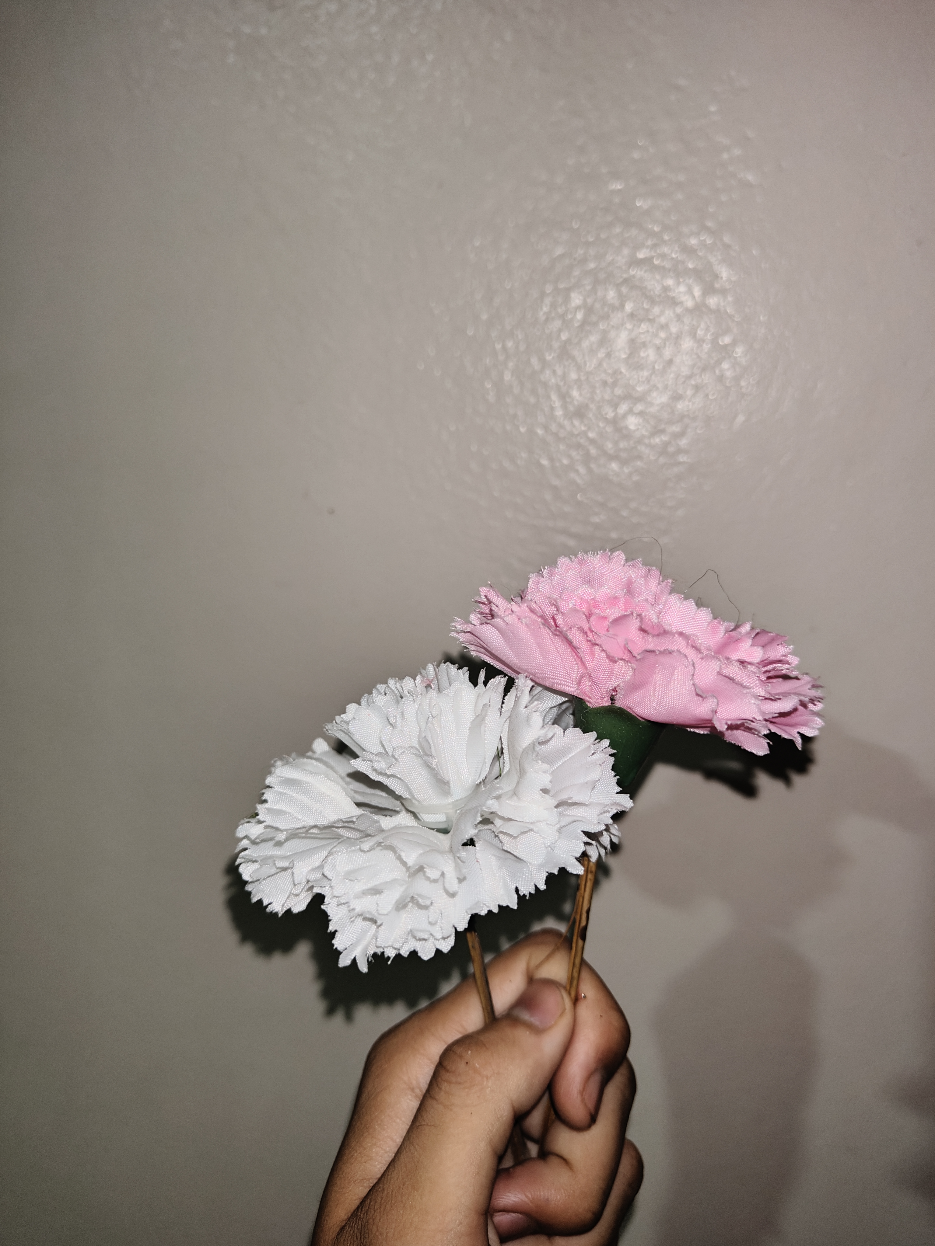 A hand holding two artificial flowers, one white and one pink, against a plain wall background.