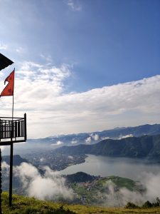 

From a wooden balcony with a red flag, a panoramic view spans Pokhara’s valley, Lake Phewa, the city, and rolling mountains under a partly cloudy, fog-kissed sky.