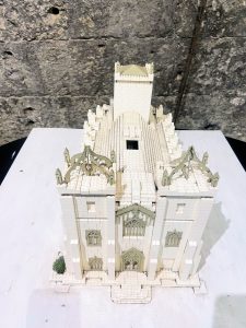 A detailed, white LEGO model of an ornate Gothic-style building with spires and arches is displayed on a white surface against a dark, rough stone wall.