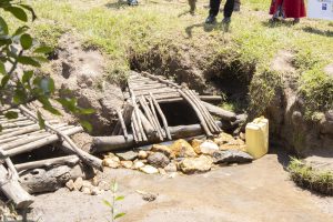 A small stream flows through a rocky area, where a well made of wooden logs and branches crossing over it. Beside the stream, there are some scattered stones and a yellow water container resting on the ground.