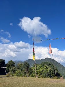 A lush green mountain landscape under a bright blue sky with white, fluffy clouds. Two flags are mounted on poles in the foreground: a tall vertical string of colorful Buddhist prayer flags on the left and a single coral-colored flag on the right.