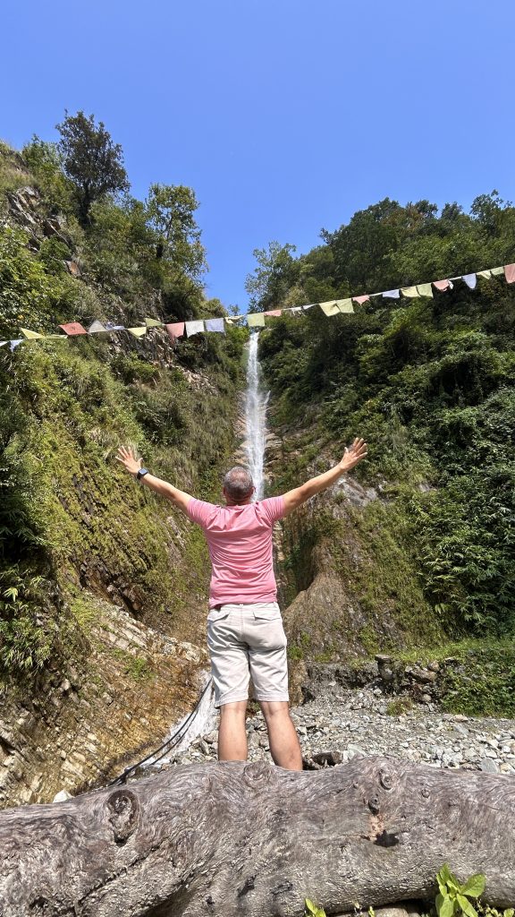 A person stands with arms outstretched in front of a waterfall surrounded by lush greenery.