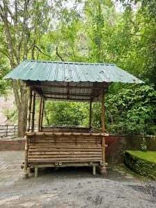 A small resting hut made with bamboo poles and a green metal roof, surrounded by lush trees near Thusharagiri Falls, Kozhikode, Kerala. 