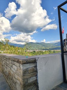 A scenic view showcasing a partially stone and concrete wall in the foreground, with lush green mountains and a town visible in the background. 