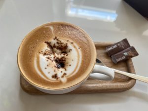 A close-up of a cappuccino with foam art in a white mug, resting on a small wooden tray with a spoon and two brown sugar packets.