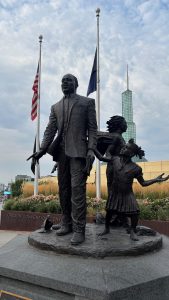 A sculpture of a man, a child, and a woman.  Behind them are two flats and the tower from a building. The child appears to be pleading with the man.