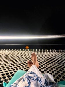 A person's bare feet are visible as they relax in a hammock at night, with a bright, full moon low on the horizon.