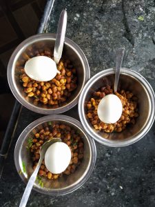 Three stainless steel bowls are arranged on a dark granite countertop, each filled with spicy roasted chickpeas, topped with a boiled egg, and served with a spoon.