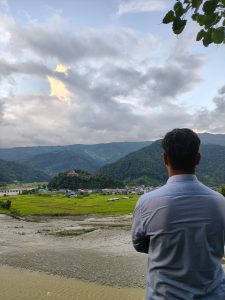 A person standing with their back to the camera, overlooking a scenic landscape featuring rolling green hills, a riverbank, and a small village with colorful houses