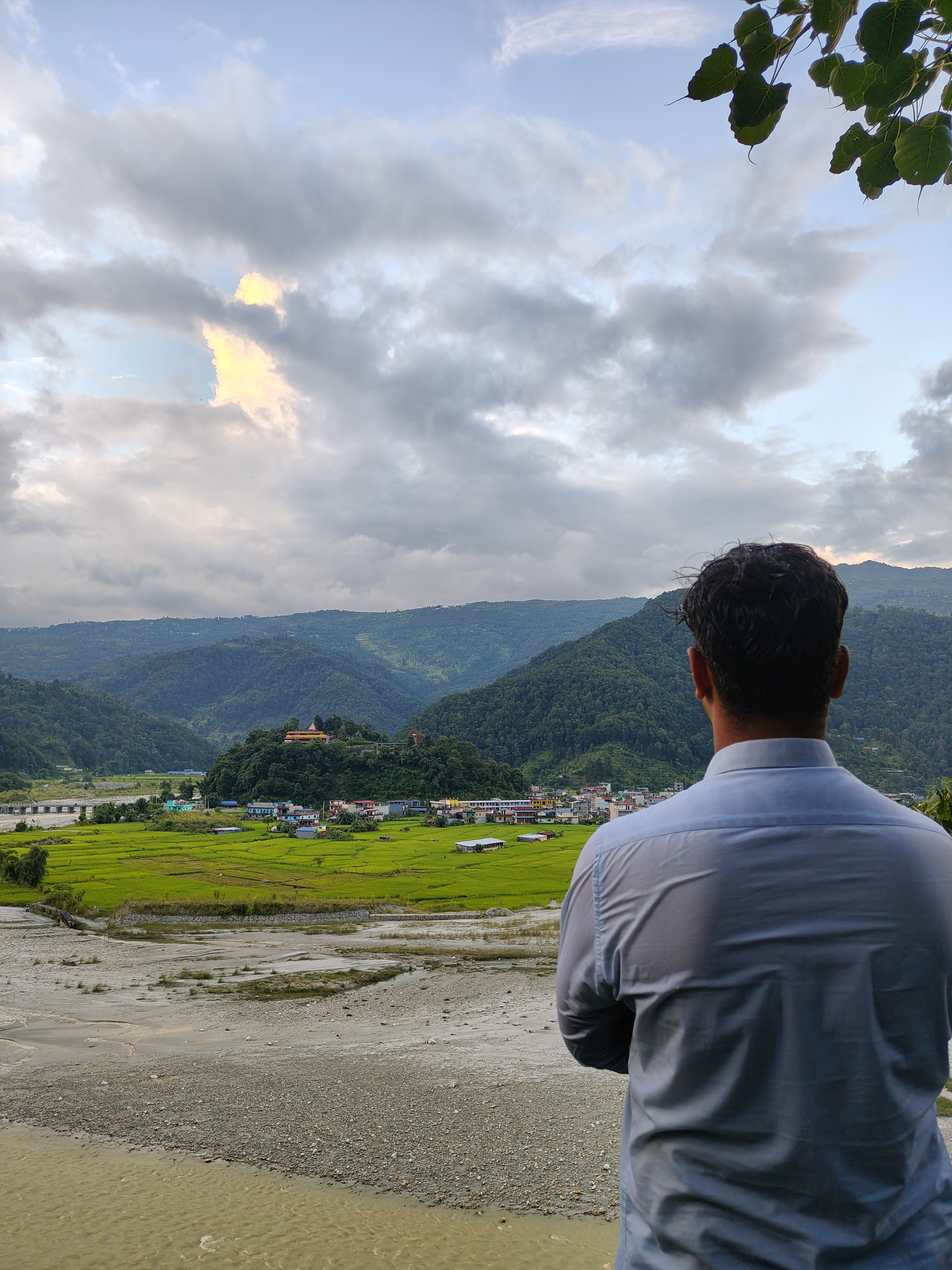 A person standing with their back to the camera, overlooking a scenic landscape featuring rolling green hills, a riverbank, and a small village with colorful houses