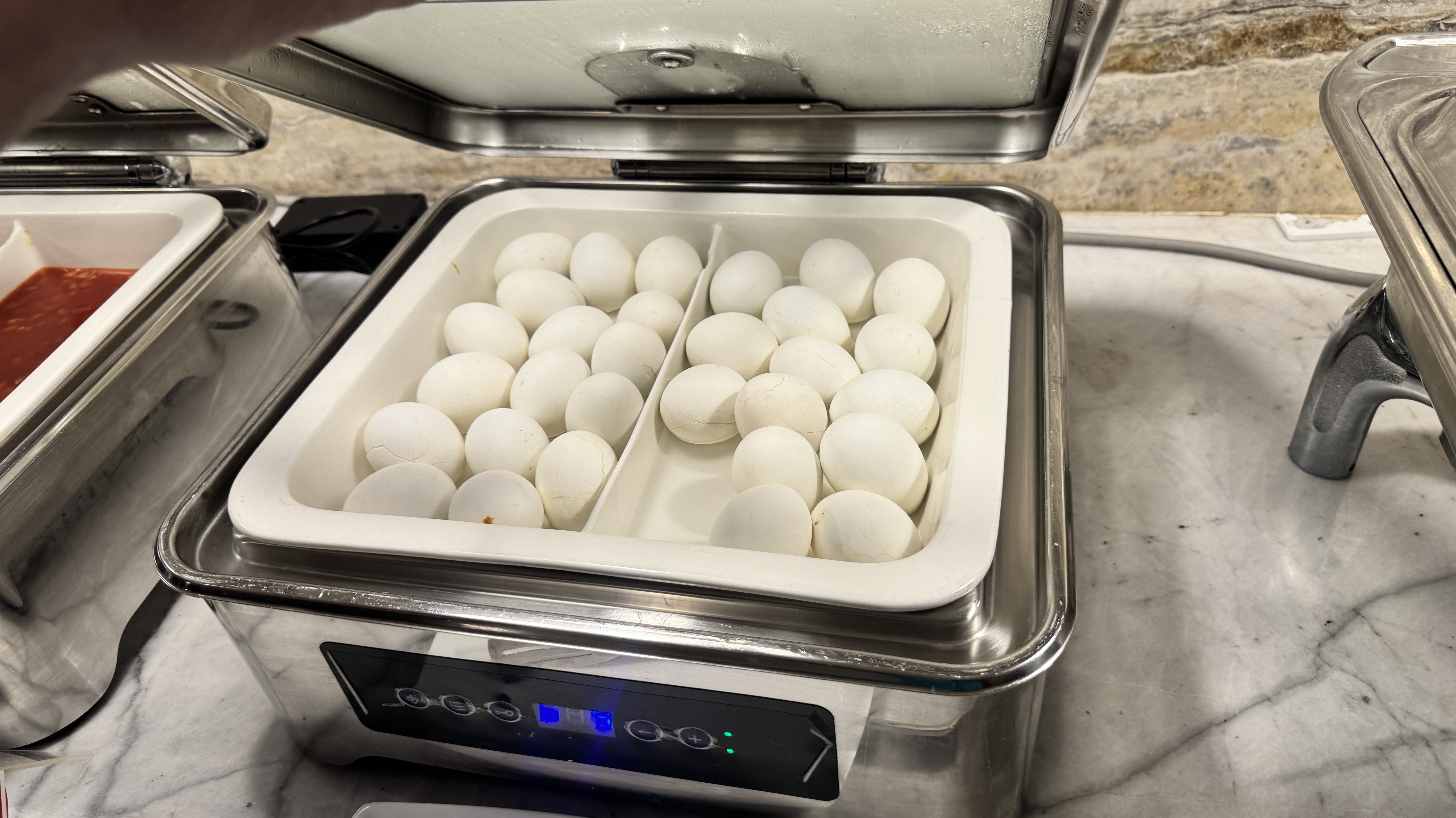 The partially lifted lid of a buffet warmer reveals a tray neatly filled with many whole, white hard-boiled eggs ready for breakfast.