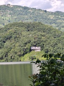 A scenic view of Begnas Lake surrounded by lush green hills. The lake reflects the greenery and a sign reading "BEGNAS LAKE" is prominently displayed on a hillside