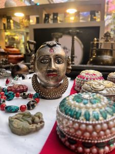 A close-up of a market stall featuring a silver metal sculpture of a face surrounded by beaded bracelets, a jade carving, and ornate jewelry boxes.