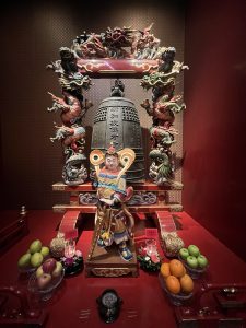 A detailed, brightly painted statue of a protective deity stands in front of a large bronze temple bell, which is framed by an elaborate wooden arch decorated with sculpted dragons.
