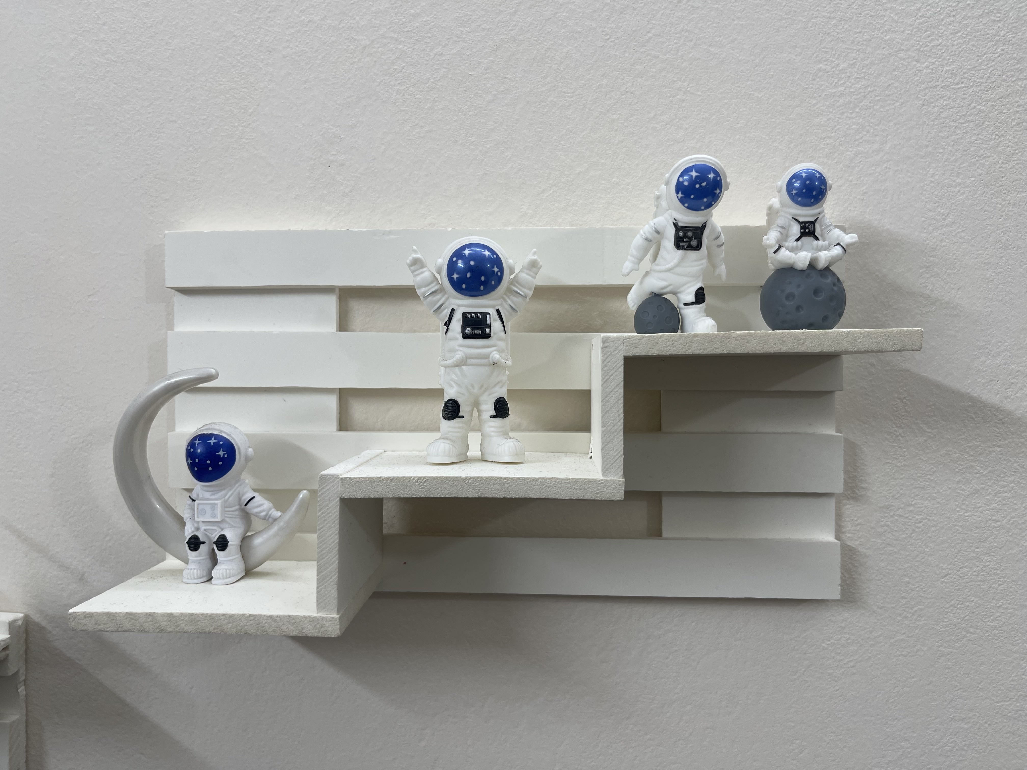 Four small astronaut figures on a white tiered shelf, one on a crescent moon and another on a grey moon.