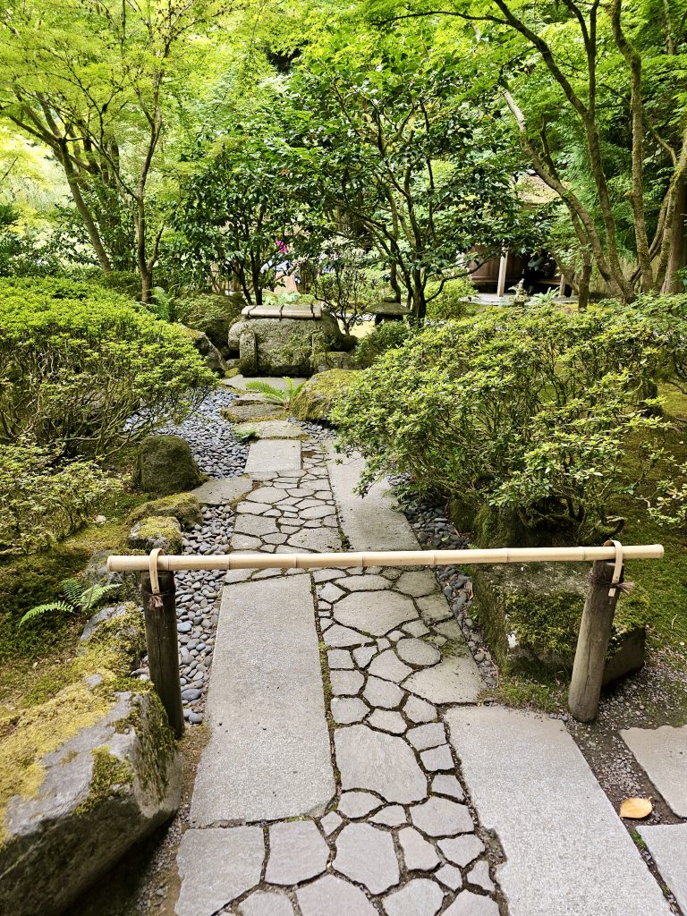 A stone path blocked by a bamboo bar leads through carefully arranged plants and rocks in the Portland Japanese Garden, creating a serene, peaceful atmosphere.