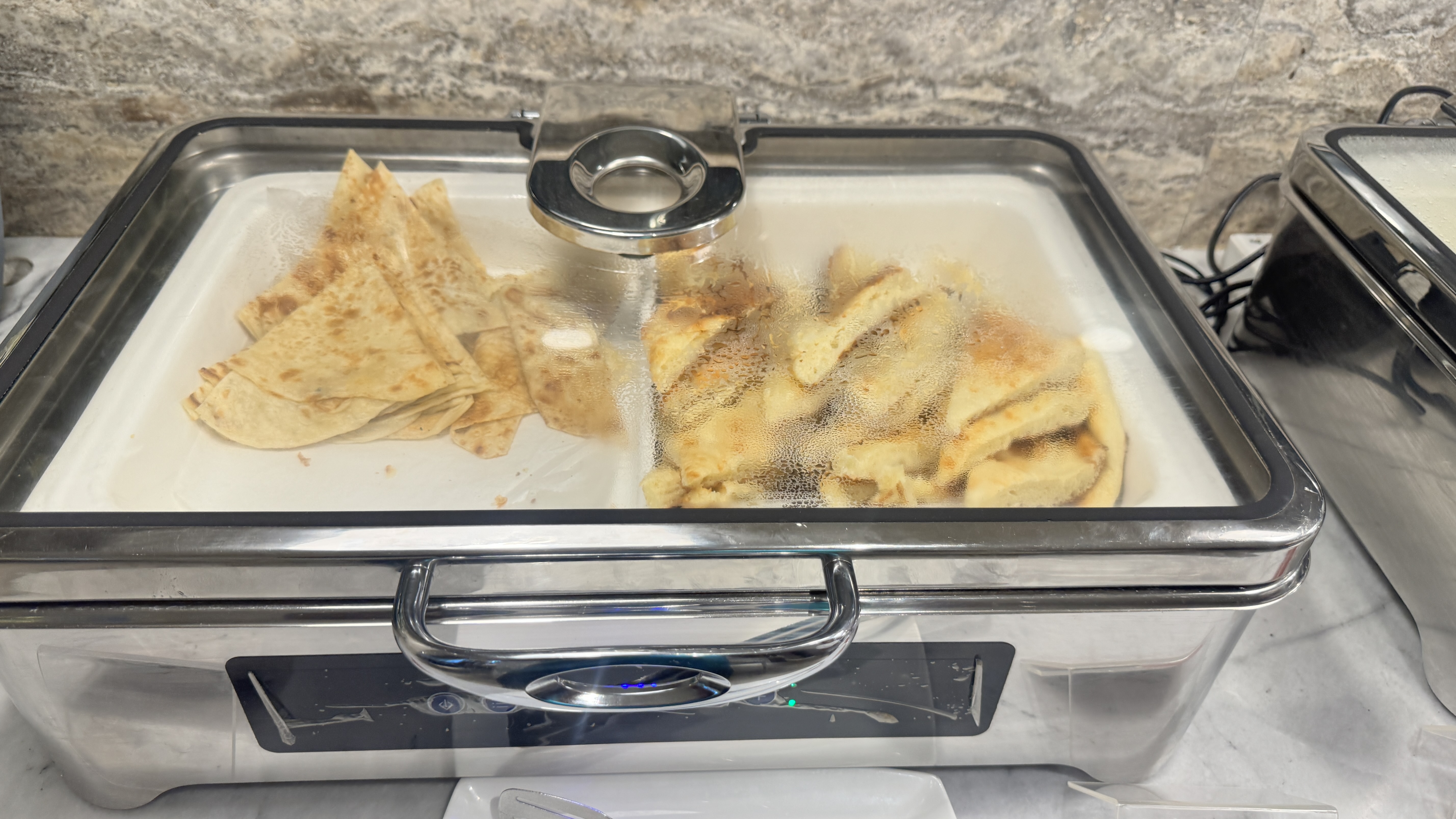 Warm naan and roti are kept in a divided tray inside a stainless steel buffet warmer, visible through the steamy glass lid.
