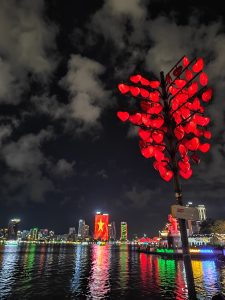 Night view of Da Nang, Vietnam, with a river reflecting colorful city lights. A tree decorated with glowing red heart-shaped lanterns stands in the foreground, while a tall building across the river is lit up with the Vietnamese flag.