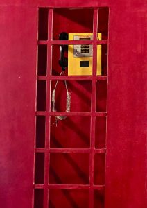 A bright yellow payphone is enclosed within a red metal grate structure, set against a deep red background.