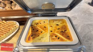 A baked English cake or pudding, sprinkled with raisins and cut into triangles, is presented in a white dish inside a buffet warmer.