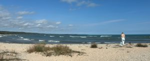A wide view with beach in the foreground, a large body of water in the middle under a blue sky.  A person in a dress walks the beach on the right.