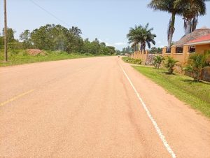 A long unpaved road lined with greenery and a low orange stone wall, captured from the Railway Station in Lira city.