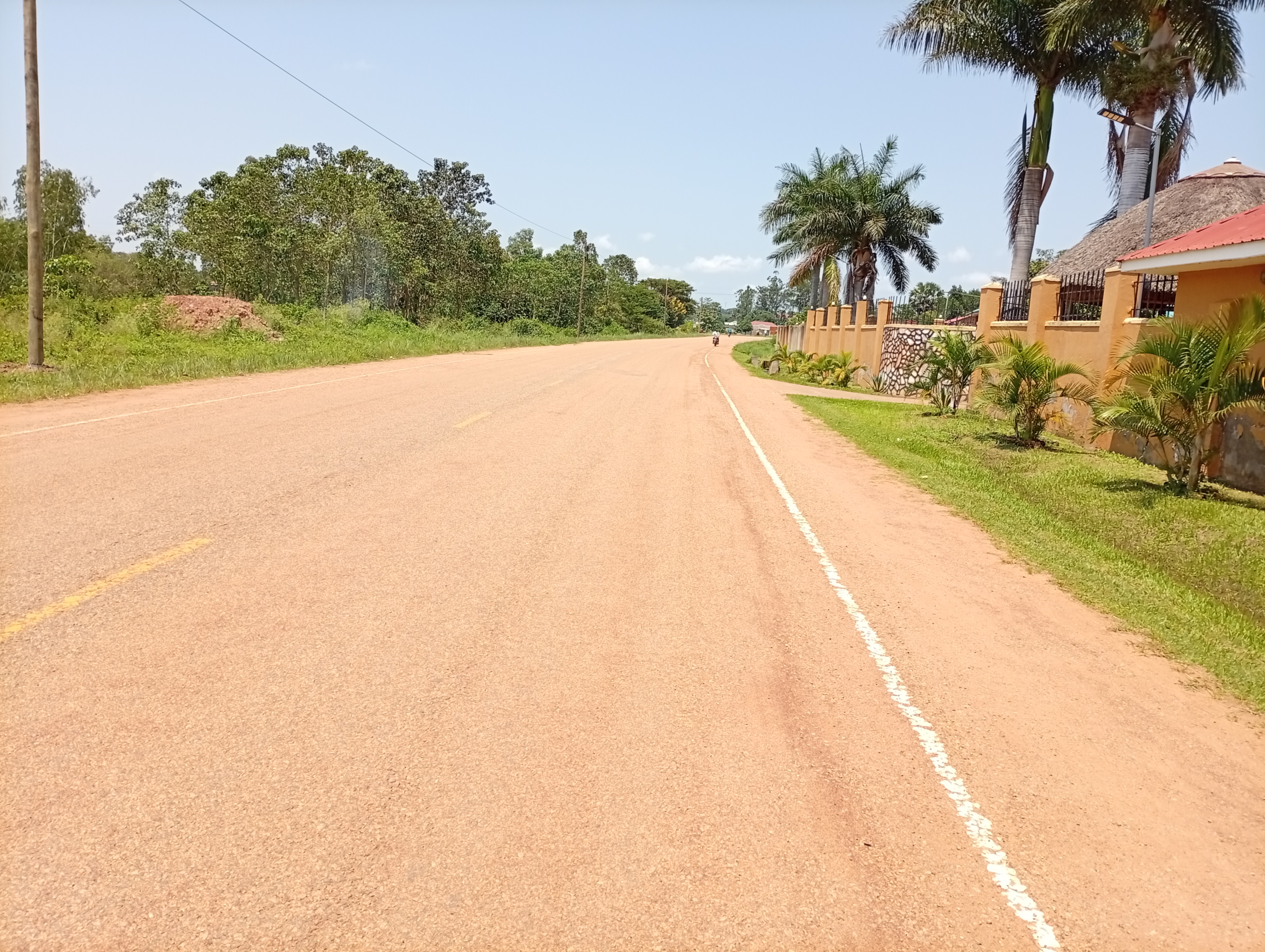 A long unpaved road lined with greenery and a low orange stone wall, captured from the Railway Station in Lira city.