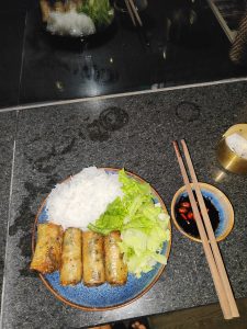A blue plate holds four crispy spring rolls, rice noodles, and fresh lettuce, served with a bowl of soy sauce with red chili and wooden chopsticks in Hanoi, Vietnam.