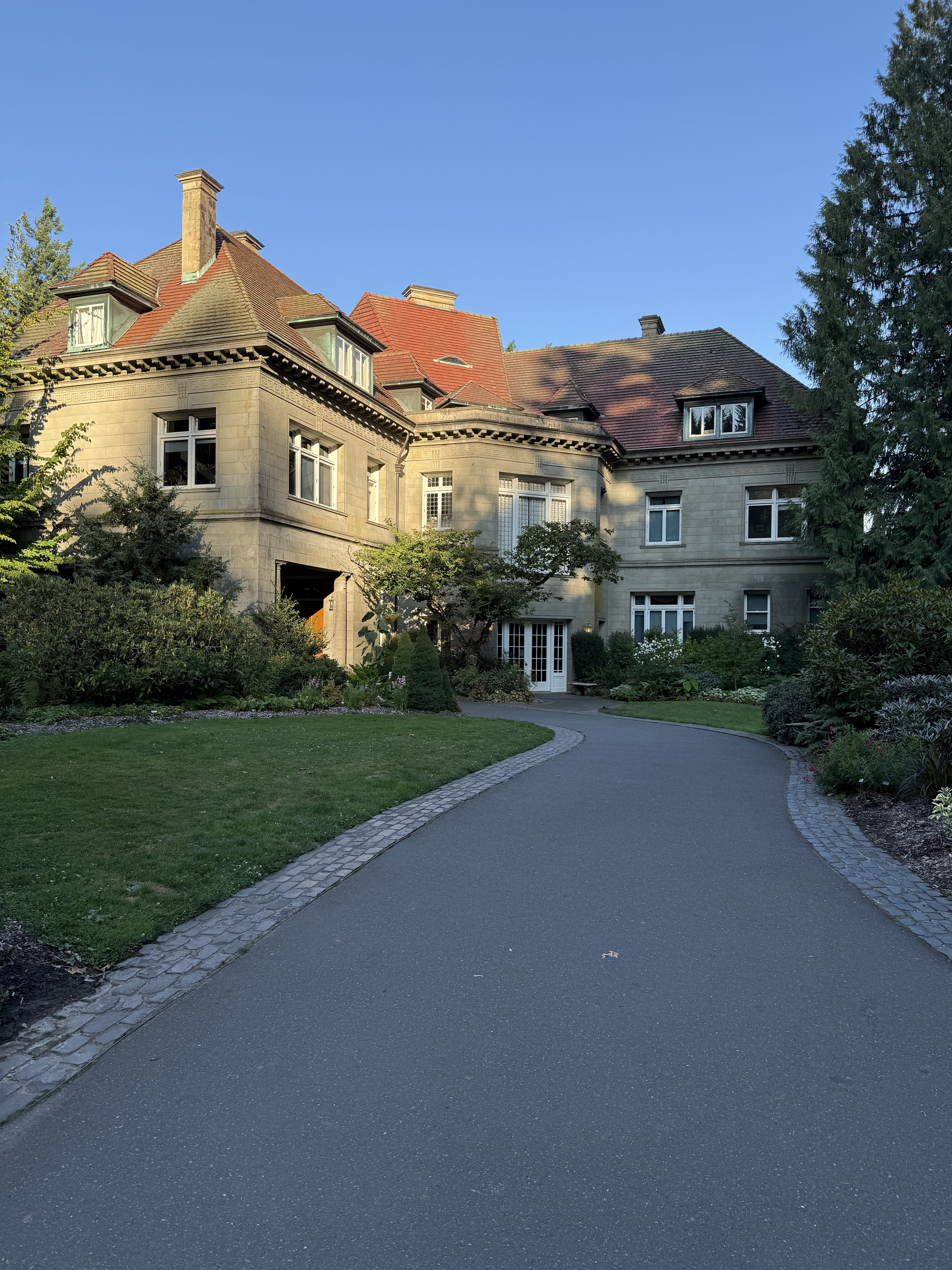A beautiful view of the Pittock Mansion in Portland, Oregon, captured in the evening light. The historic building, trees, and curved driveway are clearly visible.