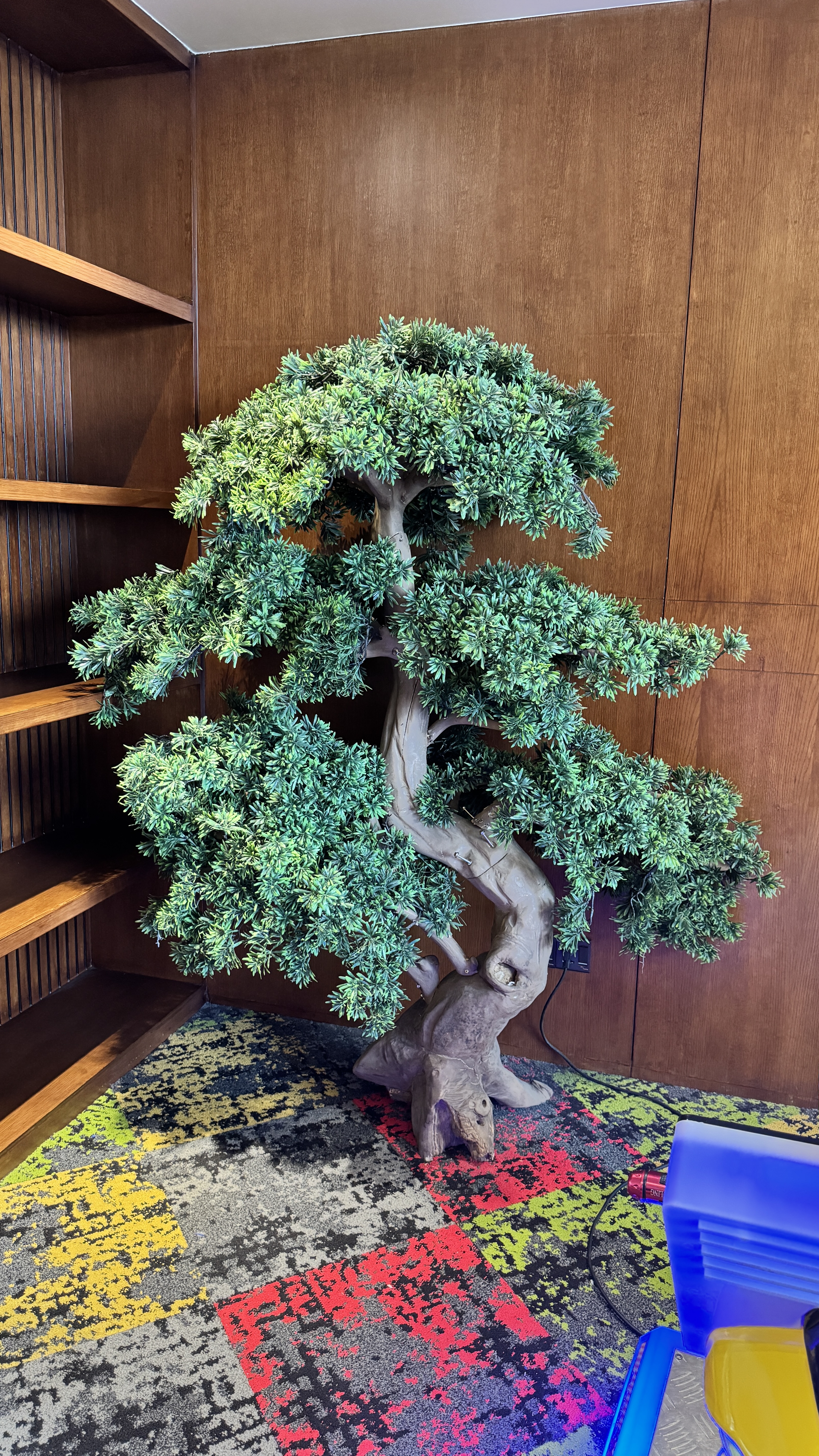 A large, artificial bonsai-style tree with a gnarled trunk and dense green leaves stands in a wood-paneled room on a colorful carpet.