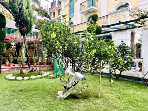 A shiny, silver abstract sculpture of a person sitting is placed in a well-maintained green lawn surrounded by trees with yellow fruit, in front of a yellow building.
